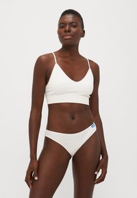 Fehér bralette vékony pántokkal és V-kivágással; hozzáillő fehér bikini alsó; sima textúra, minimalista dizájn; kék címke kiemelések.