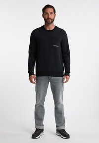 Felpa nera con tasca frontale e logo bianco. Indossata con jeans grigi chiari e sneakers nere con accenti rossi.