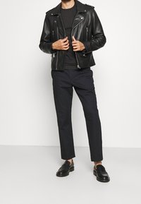 Schwarze Lederbikerjacke mit silbernen Reißverschlüssen, getragen über einem schwarzen Hemd; kombiniert mit dunklen Hosen und schwarzen Slippern. Glatte Textur insgesamt.