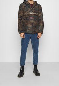 Maastokuvioitu anorakki tummanvihreissä ja ruskeissa sävyissä, jossa etutasku, logopatchi ja säädettävä huppu; yhdistetty sinisiin farkkuihin.