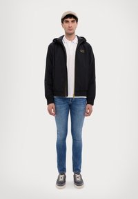 Black zip-up jacket with a hood, gold accents, worn over a white polo shirt, paired with blue jeans and dark sneakers. Beige cap on head.