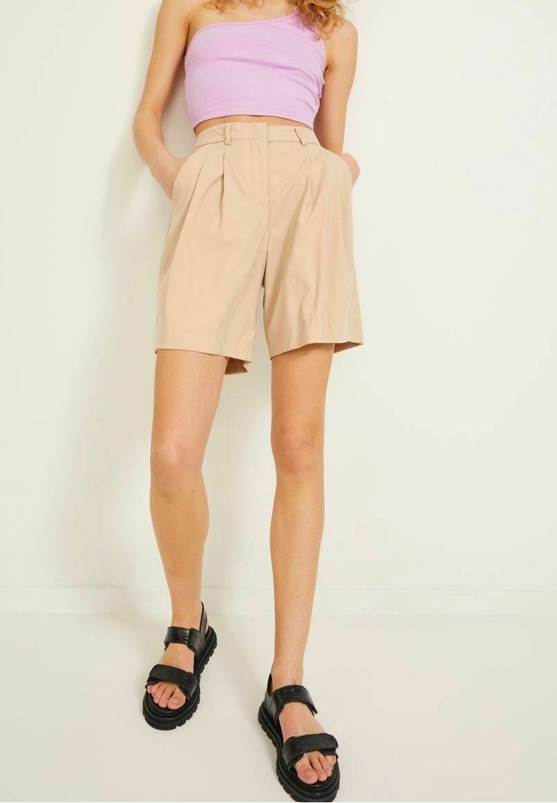 Béžové plisované šortky s bočními kapsami, zkombinované s levandulovým žebrovaným crop topem. Černé sandály s tlustou podrážkou doplňují outfit.