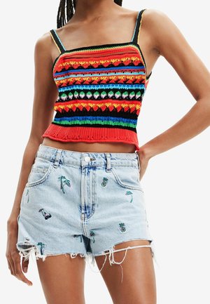 Frau trägt ein buntes, gestricktes Crop-Top mit gemusterten Streifen und hellblaue Jeans-Shorts, bestickt mit Ananas und Palmen.