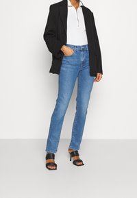 Blaue gerade geschnittene Jeans mit einer mittelhohen Taille, kombiniert mit einem weißen Reißverschlussoberteil und einem schwarzen oversized Blazer. Schwarze Absatzsandalen runden den Look ab.