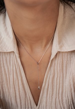 Die silberne Anhänger-Kette verfügt über eine dünne Kette, an der ein tropfenförmiger Charm hängt, der auf einem strukturierten hellen Stoff in der Nähe des Kragens ruht.