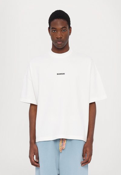 T-shirt blanc surdimensionné à manches courtes, col rond, avec "BONSAI" imprimé en noir sur le devant. Associé à un pantalon bleu clair.