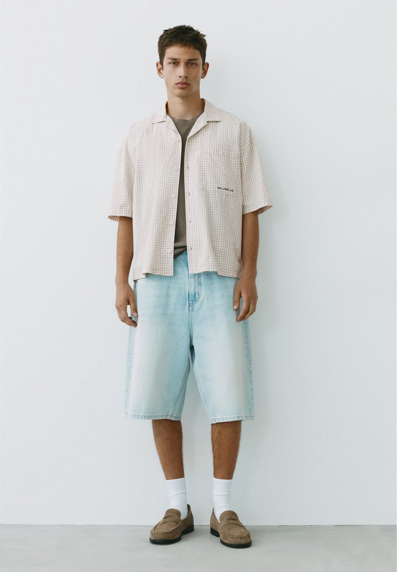 Jeune homme debout contre un mur blanc portant une chemise beige à carreaux à manches courtes, un t-shirt gris, un short en denim clair, des chaussettes blanches et des chaussures marron.