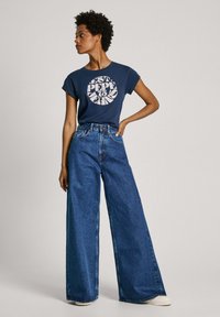 Camiseta de algodón azul marino con estampado gráfico blanco, combinada con pantalones vaqueros de pierna ancha y cintura alta de mezclilla azul. Zapatillas de colores claros.
