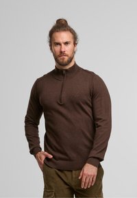 Hnědý svetr se zipem u krku, vyrobený z měkké látky. Má ribbovanou oblast u krku, rukávy a lem. Modelka stojí s rukou na boku a ukazuje design a střih.