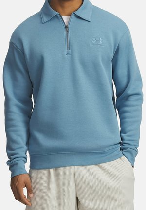Homme portant un sweat-shirt bleu clair à fermeture éclair quart, manches longues et shorts blancs texturés, mains reposant près des poches.
