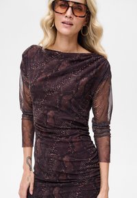 Brązowa sukienka w zwierzęcy wzór z długimi, przezroczystymi rękawami, marszczeniami i dopasowanym fasonem. Posiada dekolt typu łódka i miękką fakturę.