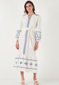White long-sleeved dress with blue embroidered accents, a deep V-neck, a belt at the waist, and a ruffled hem, made from lightweight fabric.