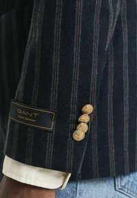 Blazer a righe navy con pattern rossi e verdi. Presenta bottoni dorati sui polsini, tessuto testurizzato e un'etichetta GANT sulla manica.