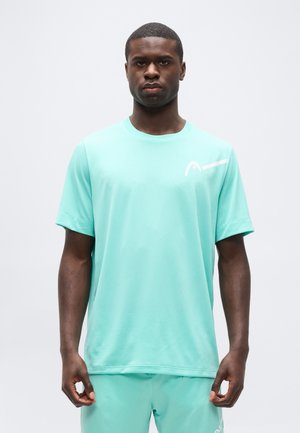 Man draagt een licht turquoise sportshirt en bijpassende korte broek, staat recht vooruit met een neutrale uitdrukking tegen een effen achtergrond.