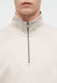 Nahaufnahme einer Person, die einen beigen Half-Zip-Pullover trägt, dessen Reißverschluss bis zum hohen Kragen vollständig geschlossen ist.