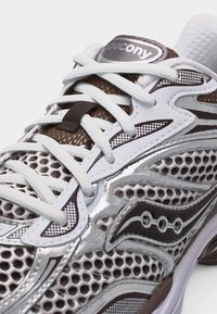 Λευκό και καφέ αθλητικό παπούτσι Saucony με αεριζόμενο δίχτυ, λευκά κορδόνια και λογότυπο της μάρκας στο πλάι και στη γλώσσα.