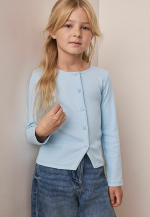Młoda dziewczyna z długimi blond włosami nosi niebieski zapinany sweter i dżinsową spódnicę, stojąc przy jasnej ścianie.