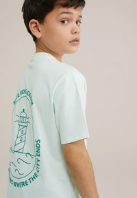 Miļļzāles zaļas īsās piedurknes T-krekls ar bāka grafiku aizmugurē, kurā ir teksts "BLUE RIDGE DENIM" un viļņu detaļas. Mīksts kokvilnas materiāls.