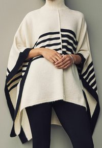 Poncho de punto en marfil con rayas horizontales negras, que presenta un cuello alto y un dobladillo asimétrico, textura suave y mangas sueltas.