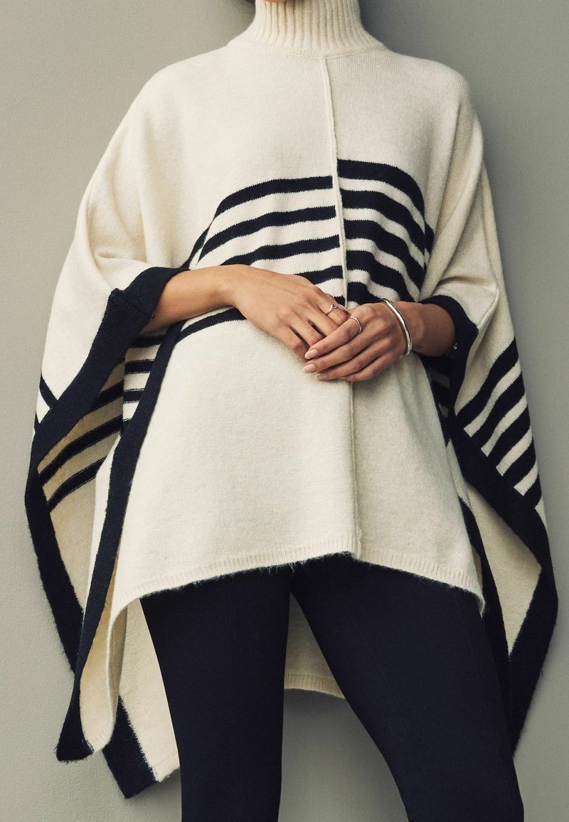 Poncho de punto en marfil con rayas horizontales negras, que presenta un cuello alto y un dobladillo asimétrico, textura suave y mangas sueltas.
