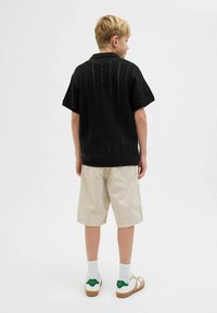 Chemise noire en tricot à manches courtes avec un col, associée à un short beige clair jusqu'aux genoux et des chaussettes blanches, accentuée par des baskets vertes et blanches.