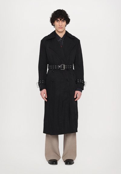 Sort trenchcoat med tekstureret finish, dobbeltbrystet design, brede kraver og sølvdetaljer. Sammen med brede beige bukser.