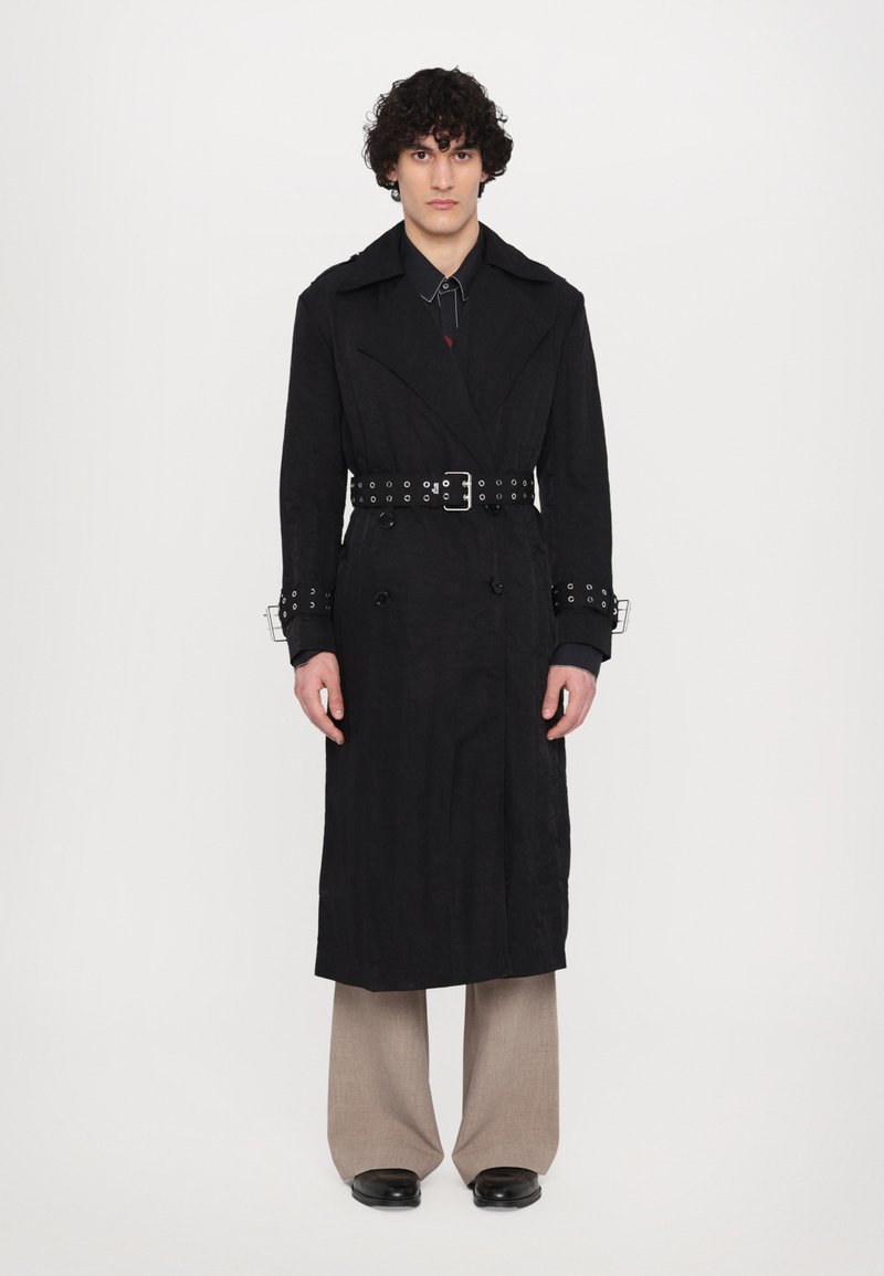 Schwarzer Trenchcoat mit strukturiertem Finish, doppelt geknöpftem Design, breiten Revers und silbernen Akzenten. Kombiniert mit weiten beigen Hosen.