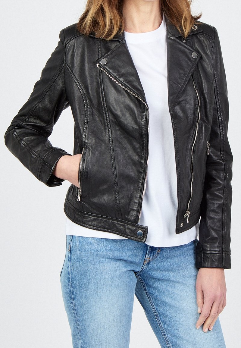 Personne portant une veste de motard en cuir noir sur un t-shirt blanc et un jean bleu clair, avec une main dans la poche de la veste.