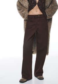Bruine broek met wijde pijpen, gemaakt van zwaar katoen met een platte voorkant, voorzien van riemlussen en een strik in de taille. Draagt een donzige jas erbij.