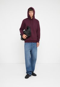 Bordeaux hoodie met trekkoordjes, zwarte tas, blauwe denim jeans en zwarte schoenen. De stof lijkt zacht en het ontwerp is casual en ontspannen.
