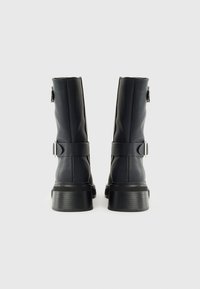 Botas negras de cuero liso hasta la rodilla con un acabado mate, que cuentan con un detalle de correa y hebilla, y una suela gruesa y texturizada.