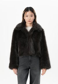 Chaqueta corta de piel sintética marrón con un amplio collar, mangas largas y una textura suave. Combinada con jeans oscuros de tiro alto.