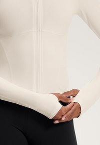 Beige pitkähihaisella paidalla, jossa on täysi vetoketju ja slim fit -muotoilu, mukana peukalolenkit hihoissa. Sujuva tekstuuri, yhdistettynä mustiin leggingseihin.