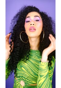 Model with long, curly black hair wearing a fitted green and yellow marbled long-sleeve top, large gold hoop earrings, against a purple background.