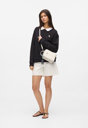 Joven mujer de pie con sudadera negra con cuello blanco, pantalones cortos blancos, sandalias negras y un bolso blanco cruzado sobre un fondo liso.