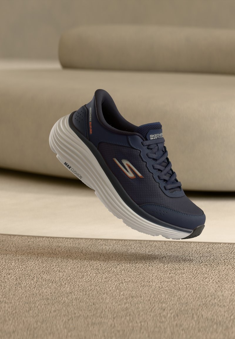 Ναυτικό μπλε αθλητικό παπούτσι Skechers με λευκή χοντρή σόλα, σχεδιασμένο για μέγιστη άνεση Max Cushion, τοποθετημένο πάνω σε μπεζ χαλί και φόντο καναπέ.