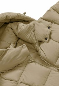 Beige pufferjack met een geborduurd ontwerp en zichtbare stiksels. Ritsluiting met drukknoopjes en textuurstof. Zacht materiaal zichtbaar.