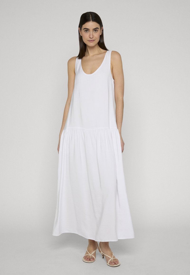 Kvinde med glat mørkt hår iført en hvid maxikjole uden ærmer og beige sandaler med hæl og remme, stående mod en ensfarvet baggrund.