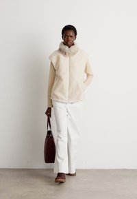 Gilet in pelliccia sintetica beige con colletto alto, indossato sopra una maglia a maniche lunghe. Abbinato a pantaloni bianchi e una borsa marrone, con scarpe basse.