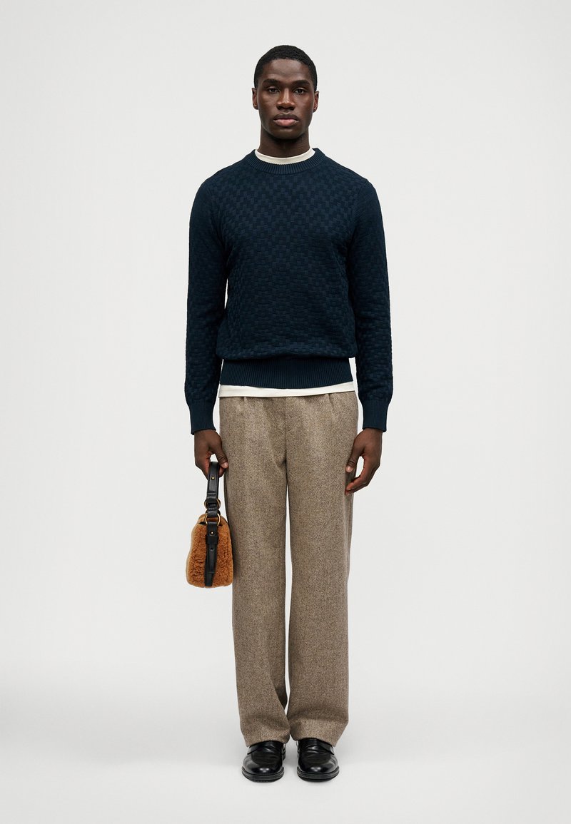 Uomo che indossa un maglione testurizzato blu navy, pantaloni beige a gamba larga, scarpe nere, mentre tiene in mano una piccola borsa di pelliccia marrone su sfondo neutro.