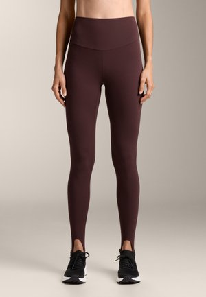 Burgundowe legginsy z wysokim stanem, wykonane z gładkiego, elastycznego materiału, mają elegancki design z bocznymi rozcięciami na kostkach. Połączone z czarnymi butami sportowymi.