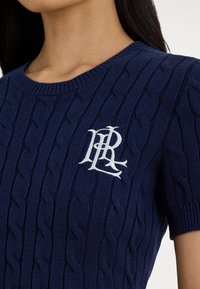 Moteris dėvi tamsiai mėlyną trumpomis rankovėmis megztą megztinį su baltu siuvinėtu monograma ant krūtinės.