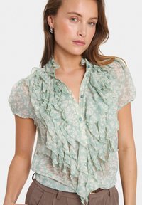 Ljust grön, genomskinlig blus med volang framtill, korta ärmar och blommönster. Har en krage och knappar framtill, i kombination med byxor.