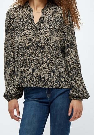 Femme portant un chemisier à manches longues beige avec motif abstrait noir, col en V, et un jean bleu, debout devant un fond uni.