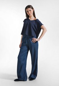 Námořnicky modrý krátký top se širokými rukávy, spárovaný s volnými dlouhými tmavými džínovými kalhotami s nastavitelným pasem na šňůrku, hladkou texturou a bočními rozparky.