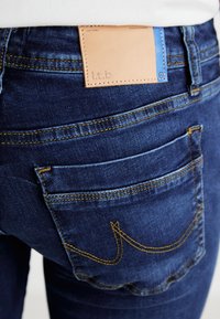 Jean bleu foncé avec patch en cuir au dos arborant un logo en relief, surpiqûres jaunes, et deux poches arrière avec des coutures incurvées.