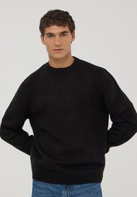 Czarny dzianinowy sweter z okrągłym dekoltem i ściągaczami na rękawach, w połączeniu z niebieskimi dżinsami. Tkanina wydaje się miękka i teksturowana.