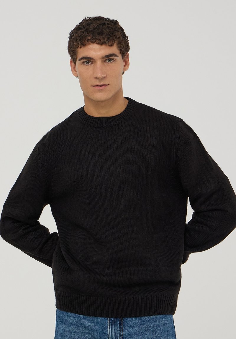 Czarny dzianinowy sweter z okrągłym dekoltem i ściągaczami na rękawach, w połączeniu z niebieskimi dżinsami. Tkanina wydaje się miękka i teksturowana.