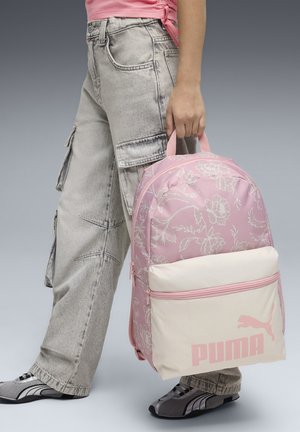 Mochila de patrones florales en tonos rosas con base color crema, que cuenta con un bolsillo frontal y un logo rosa. Sostenida por una persona con pantalones cargo grises.
