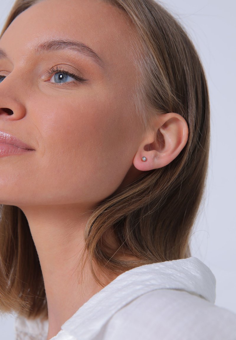 Boucles d'oreilles rondes avec une pierre précieuse transparente. Le modèle a une coiffure courte et ébouriffée et porte une chemise blanche à col. Texture de peau lisse.